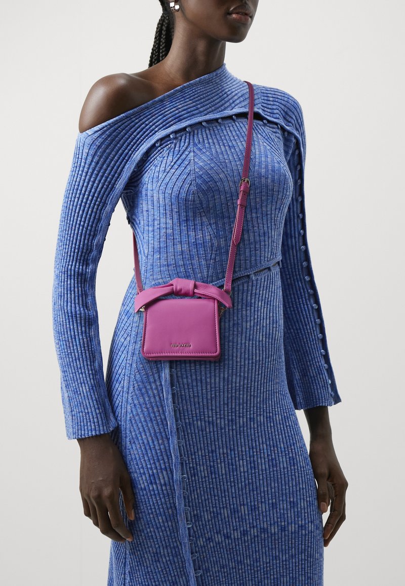Donna che indossa un vestito blu a costine con spalle scoperte e una piccola borsa a tracolla viola con un dettaglio a fiocco sul davanti.