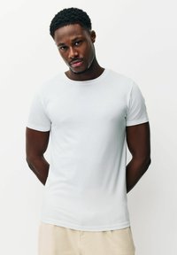 Mladý černoch s krátkými kudrnatými vlasy, oblečený v jednoduchém bílém tričku s krátkým rukávem a béžových kalhotách, stojící na bílom pozadí.