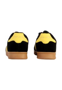 Zapatillas de ante negras y amarillas con suela de goma, que presentan un acento amarillo curvo en el talón y tiras amarillas en los lados.