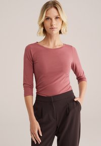 Top a maniche lunghe di colore rosa realizzato in un morbido tessuto elasticizzato, con scollatura rotonda e silouette aderente, abbinato a pantaloni scuri a vita alta.