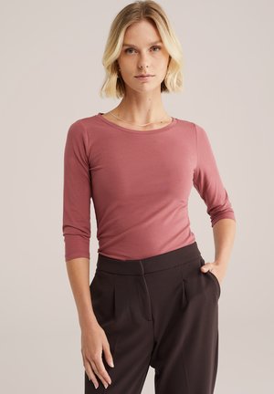 Top a maniche lunghe di colore rosa realizzato in un morbido tessuto elasticizzato, con scollatura rotonda e silouette aderente, abbinato a pantaloni scuri a vita alta.
