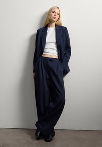Jeune femme aux longs cheveux blonds portant un blazer oversize navy et un pantalon large, un top blanc court, des chaussures noires, s'appuyant contre un mur blanc.