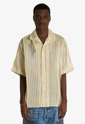 Uomo che indossa una camicia beige a maniche corte con bottoni e righe verticali e jeans blu, rivolto in avanti con espressione neutra.