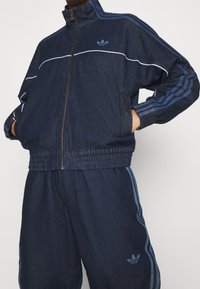 Person som bär en mörkblå Adidas träningsoverall med tre ränder på ärmarna och byxorna, med händerna i jackfickorna, stående mot en enfärgad bakgrund.