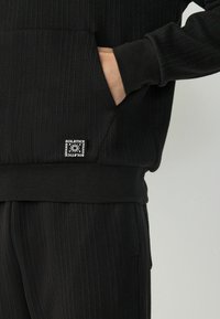 Hanorac negru cu nervuri, dotat cu o bătaie frontală, având un patch cu logo. Materialul pare moale și texturat, cu o talie și manșete ajustate.