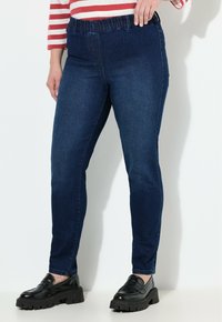 Žena v tmavě modrých slim-fit džínech s elastickým pasem, pruhované long-sleeve tričko a černé robustní mokasíny, stojící proti bílé zdi.