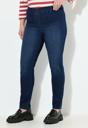 Frau in dunkelblauen Slim-Fit-Jeans mit elastischem Bund, gestreiftem Langarmshirt und schwarzen klobigen Slippers, die an einer weißen Wand steht.