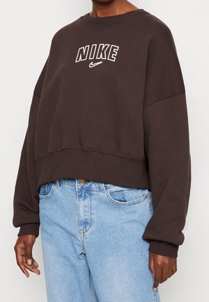 Persoon draagt een donkerbruine Nike-sweatshirt met logo over de borst en lichtblauwe hoge taille jeans, staand tegen een witte achtergrond.