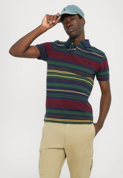 Polo cu dungi în navy, verde, burgundy cu accente galbene; mâneci scurte; materialul are o textură moale; asortat cu pantaloni cargo bej.