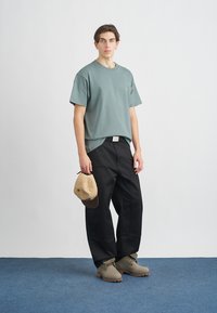 Grünes Baumwoll-T-Shirt, weiter Schnitt, schwarze Hosen mit weitem Bein, hellbraune Mütze in der Hand gehalten, graue Stiefel, minimalistisches Design mit dezenten Akzenten.