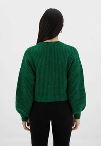 Zielony sweter w prążki o krótkim kroju i oversize'owych rękawach, wykonany z teksturowanej dzianiny. Noszony z czarnymi spodniami z wysokim stanem.