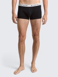 Modello maschile in piedi a piedi nudi, indossa slip neri con elastico bianco marchiato "Tom Tailor" su uno sfondo chiaro e neutro.