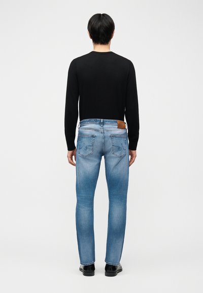 Άνδρας που φοράει μαύρο μακρυμάνικο πουκάμισο, ανοιχτό μπλε στενά τζιν και μαύρα παπούτσια, στέκεται με την πλάτη στραμμένη σε απλό ανοιχτόχρωμο φόντο.