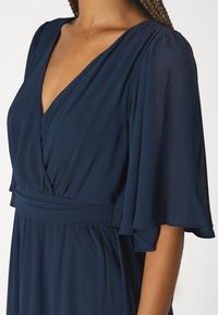 Marinblå klänning med V-ringning, svävande klockärmar och en figurnära midja. Materialet verkar lätt och slätt, med en mjuk struktur.