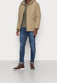 Veste matelassée beige avec capuche, avec des accents orange, associée à une chemise verte, un jean bleu foncé et des bottes marron à lacets.