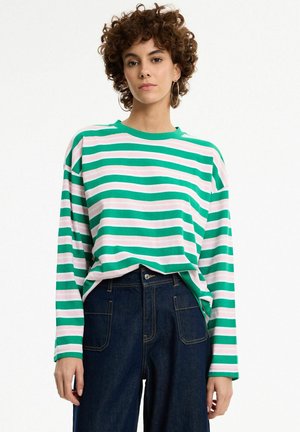 Persona joven con cabello corto y rizado que lleva una camisa de manga larga a rayas verdes y blancas y pantalones de talle alto azul oscuro sobre fondo blanco.