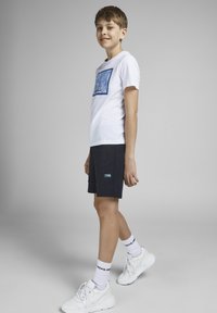 Hvit t-skjorte med blå grafisk rektangel, marineblå shorts med sømdetaljer, og hvite sneakers med teksturerte detaljer.