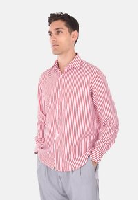Camicia a maniche lunghe a righe rosse e bianche con colletto button-down, taschino sul petto, e vestibilità ampia, abbinata a pantaloni grigi.