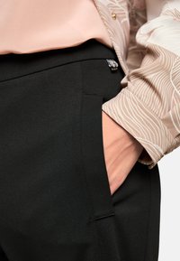 Schwarze Hose mit glatter Textur und Seitentasche, kombiniert mit einer hellrosa Bluse mit beigem wellenförmigem Muster und Knopfakzenten.