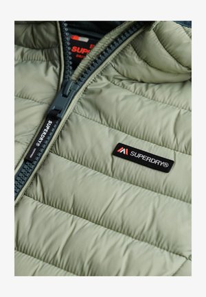 Hellegrüne isolierte Jacke mit gestepptem Design, ausgestattet mit einem dunklen Reißverschluss und einem Superdry-Logo-Patch. Reißverschlussdetails entlang der Vorderseite.