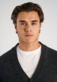 Tummanharmaa neuletakki hartiahihalla valkoisen t-paidan päällä. Neuletakki on tekstuurilla neulottu ja siinä on syvä V-pääntie.