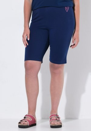 Frau trägt marineblaue knielange Shorts mit einem kleinen pinken Herz-Detail und pinke Sandalen mit Schnallen und wellenförmigen Kanten.