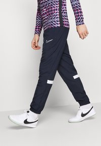 Person som går iförd svarta joggers med vita ränder, vita Nike-sneakers med svart swoosh, och en långärmad topp med lila mönster.