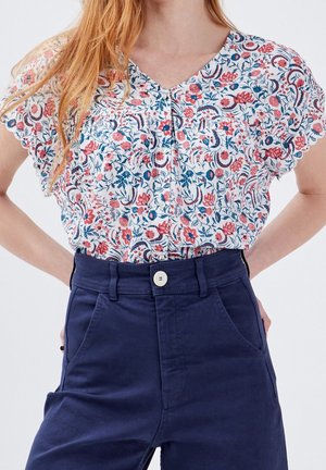 Blouse florale blanche avec imprimé rouge et bleu, décolleté en V, manches courtes et détail de boutons ; associée à un pantalon taille haute bleu marine.
