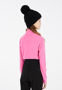 Fleece crop pulóver v jasne ružovej farbe s vysokým golierom, v kombinácii s čiernymi nohavicami s vysokým pásom. Čierna pletená čiapka s pom-pomom na vrchu.