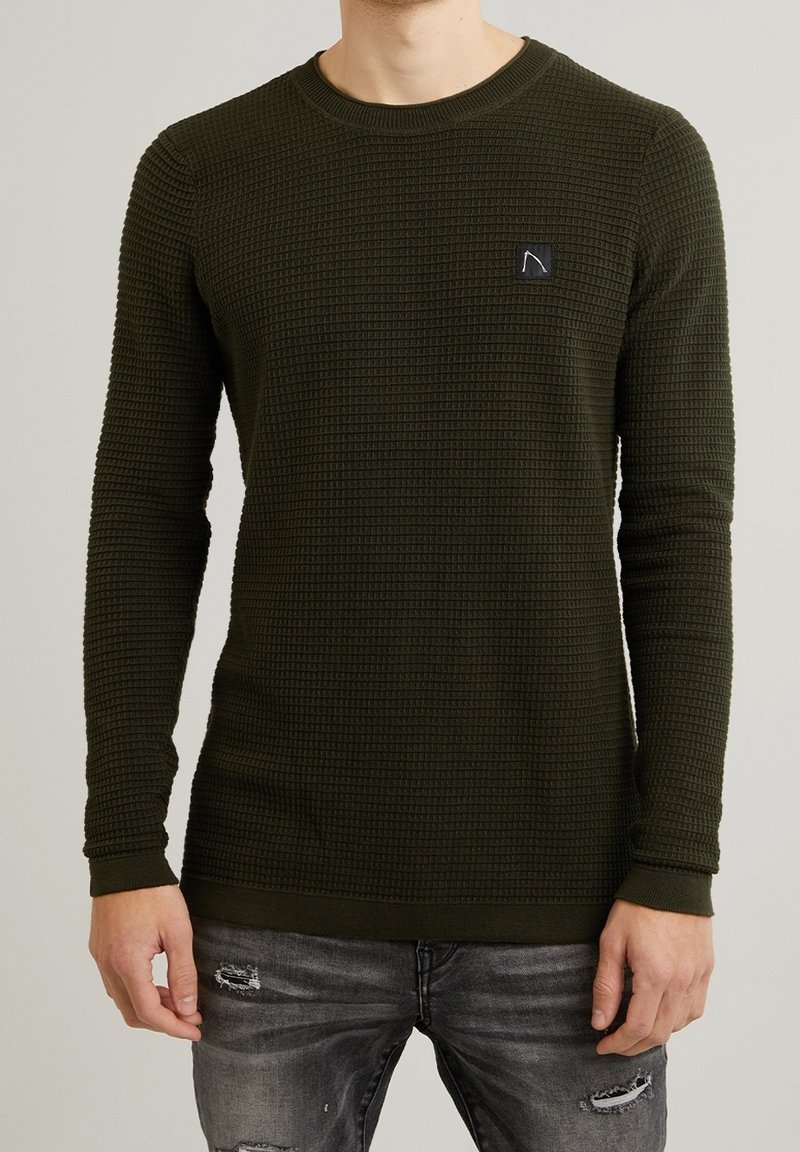 Maglione verde a maniche lunghe in maglia con un motivo testurizzato, scollatura tonda e una piccola toppa nera con logo sul petto. Indossato con jeans grigi strappati.
