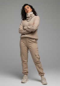 Beige ribbed sweater with a high cowl neck and chain detail, paired with beige cargo pants featuring side pockets and cuffed legs.