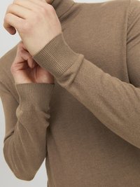 Brauner Rollkragenpullover aus weichem Strickmaterial mit gerippten Bündchen und Kragen. Nahaufnahme von Händen, die den Stoff justieren.
