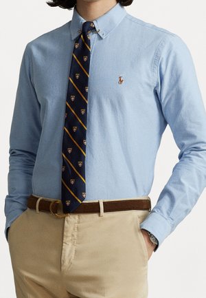 Mann trägt hellblaues Hemd mit Knöpfen und gesticktem Logo, marineblau gemusterten Schlips, beige Hose, braunen Gürtel und Armbanduhr, Hände in den Taschen.