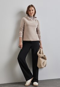 Beige hoodie met lange mouwen, gecombineerd met zwarte wijde broek. Model houdt een gestructureerde beige tas vast en draagt witte sneakers met beige accenten.