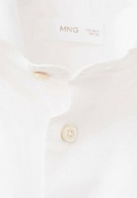 Close-up of a white collared shirt with a beige button and a partially visible MNG brand label inside the collar.