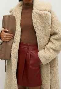 Kermansuoja faux shearling takki, ruskea sovitettu neulepaita ja burgundinpunainen nahkahame. Pitää ruskeaa nahkakukkaroa. Rakenneviljellä varustetut kankaat ja muotoiltu istuvuus.