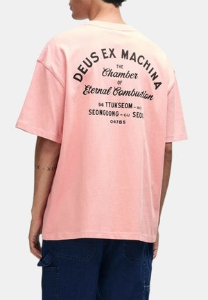 Joven que lleva una camiseta rosa holgada con texto negro que dice "Deus Ex Machina" y pantalones azul oscuro, visto desde atrás.