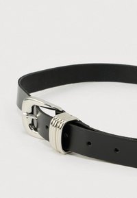 Melns ādas josta ar sudraba metāla sprādzi, kurai ir horizontālas rievas. Siksnai ir caurumi izmēra regulēšanai.