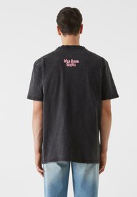 Melnas kokvilnas T-kreklis ar īsām piedurknēm, aizmugurē ar rozā grafisku tekstu "WILD ROOM SERVICE". Tieša mala, brīvs piegriezums.