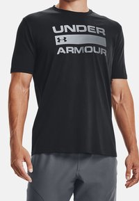 Svart t-shirt med kort ärm i lätt material som har en stor silverfärgad "Under Armour"-logotyp centrerad över bröstet.