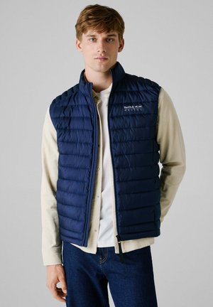 Marineblauwe gewatteerde puffer vest met hoge kraag en rits, voorzien van het Red Bull Racing-logo. Draagt over een lichtgekleurde lange mouw shirt.