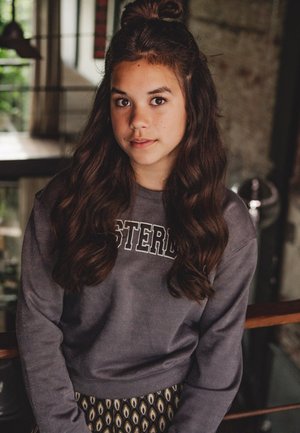 Grijze sweatshirt met de tekst "AMSTERDAM", lange golvende haren en een patroonrok met druppelmotieven, staande binnen op een houten leuning.