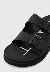 Black slide sandal with two adjustable straps, rectangular buckles, textured footbed, and contrasting white logo on the sole.