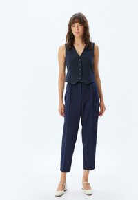 Marineblauw gestreepte mouwloze vest met een V-hals en knopen, gecombineerd met opgebroken marineblauwe broek en crèmekleurige platte schoenen.