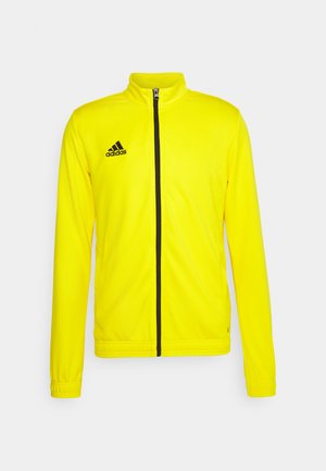 Giacca sportiva gialla brillante con zip frontale, logo Adidas nero sul petto sinistro e polsini elasticizzati. Texture liscia, design leggero.