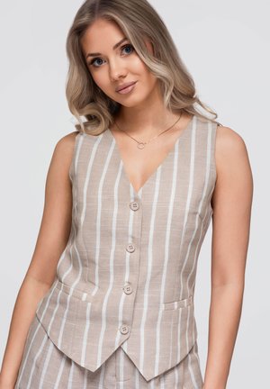 Giovane donna con capelli biondi che indossa un gilet smanicato beige e bianco a righe verticali con bottoni e pantaloni coordinati, sorridendo dolcemente.