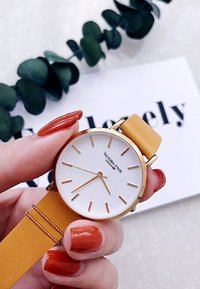 Esfera de reloj blanca redonda con marcadores de hora dorados, alojada en un bisel dorado. Presenta una correa de cuero amarilla y el nombre de la marca "Victoria Hyde."