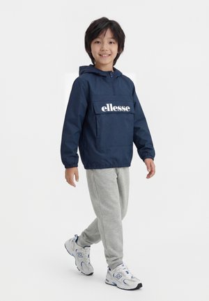 Bambino sorridente che cammina, indossando una giacca con cappuccio navy blue di ellesse, pantaloni grigi da ginnastica e scarpe da ginnastica bianche New Balance su sfondo bianco.