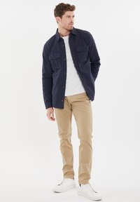 Marineblauw katoenen overhemdjack met vier zakken, knoopsluiting, gedragen over een lichtgrijze T-shirt en gecombineerd met beige broeken en witte sneakers.