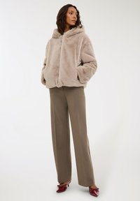 Donna con una giacca cappuccio in peluche beige, pantaloni larghi taupe e décolleté a punta rossa con fiocchi, in piedi contro uno sfondo bianco.